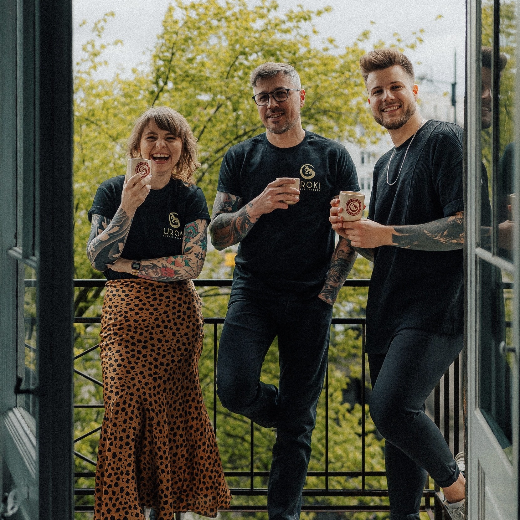 Zdjęcie trzech uśmiechniętych tatuażystow na balkonie warszawskiego studio tatuażu Uroki. Ubrani w firmową koszulkę UROKI. Wszystkie osoby z widocznymi tatuażami na rękach